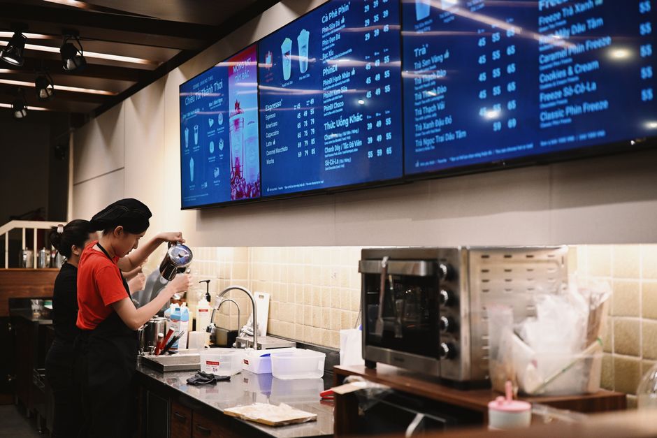 Two baristas prepare drinks in a modern coffee shop with digital menu screens.